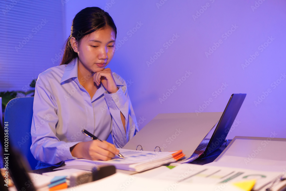 An Asian woman is working overtime at her desk in the office, busy viewing documents and her notebook. She is talking while managing tasks late at night, staying focused and productive An Asian woman is working overtime at her desk in the office, busy viewing documents and her notebook. She is talking while managing tasks late at night, staying focused and productive