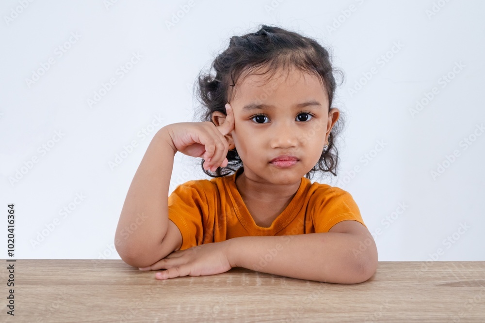 A cute little Asian girl sitting at table and posing with one hand pointing to her head. Confused, thinking, or dreaming concept. A cute little Asian girl sitting at table and posing with one hand pointing to her head. Confused, thinking, or dreaming concept.