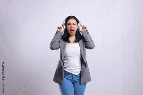 beautiful office girl screaming angry candid holding and wearing headphone headset listening music wearing grey suit isolated on white background. lifestyle, entertainment, technology concept