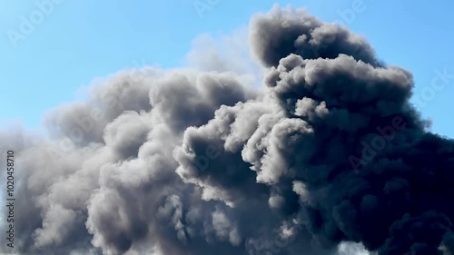 Black smoke in the sky. Huge clouds of black smoke against a blue sky. A giant fire and a high column of black smoke. Puffs of dramatic black clouds of toxic smoke moving across sky from violent fire