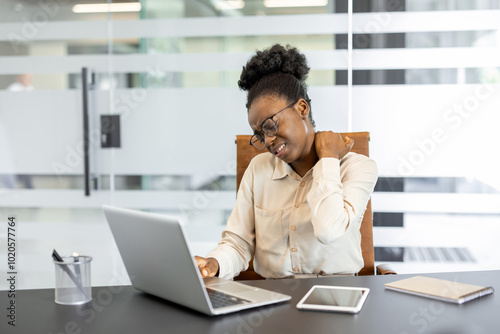 Woman at office desk using laptop and phone, experiencing neck pain. Office environment suggests long work hours, highlighting need for ergonomic workspace and stress management.