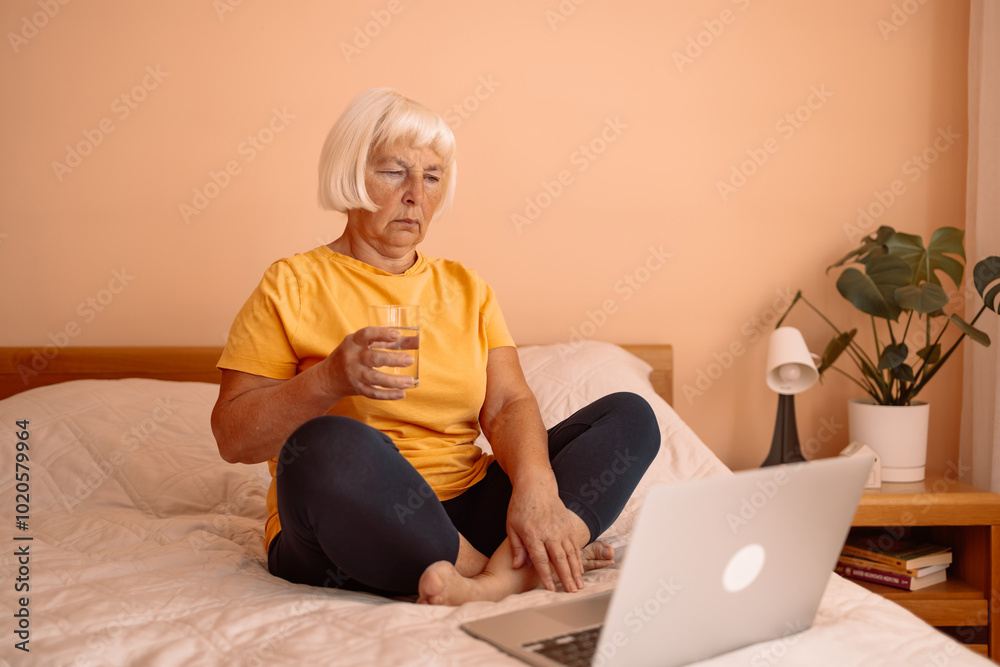 Senior 60s woman in pajamas suffering from headache sitting on bed at home bedroom, holding a glass of water. Female working on a laptop sitting on the bed in the house. health, old age and people Senior 60s woman in pajamas suffering from headache sitting on bed at home bedroom, holding a glass of water. Female working on a laptop sitting on the bed in the house. health, old age and people