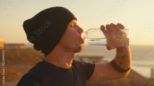 Sporty man drinking water from bottle overlooking sea and clear sky during sunrise. Young male jogger is looking away while taking break at dawn. 