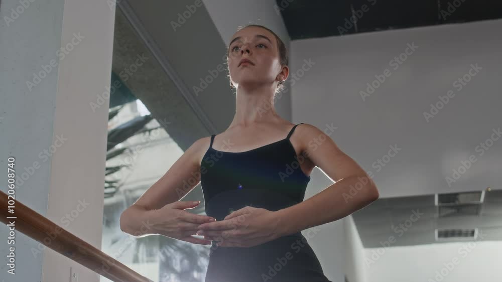 Low angle medium shot of concentrated ballet student in black leotard practicing posture at barre in sunlit studio Low angle medium shot of concentrated ballet student in black leotard practicing posture at barre in sunlit studio