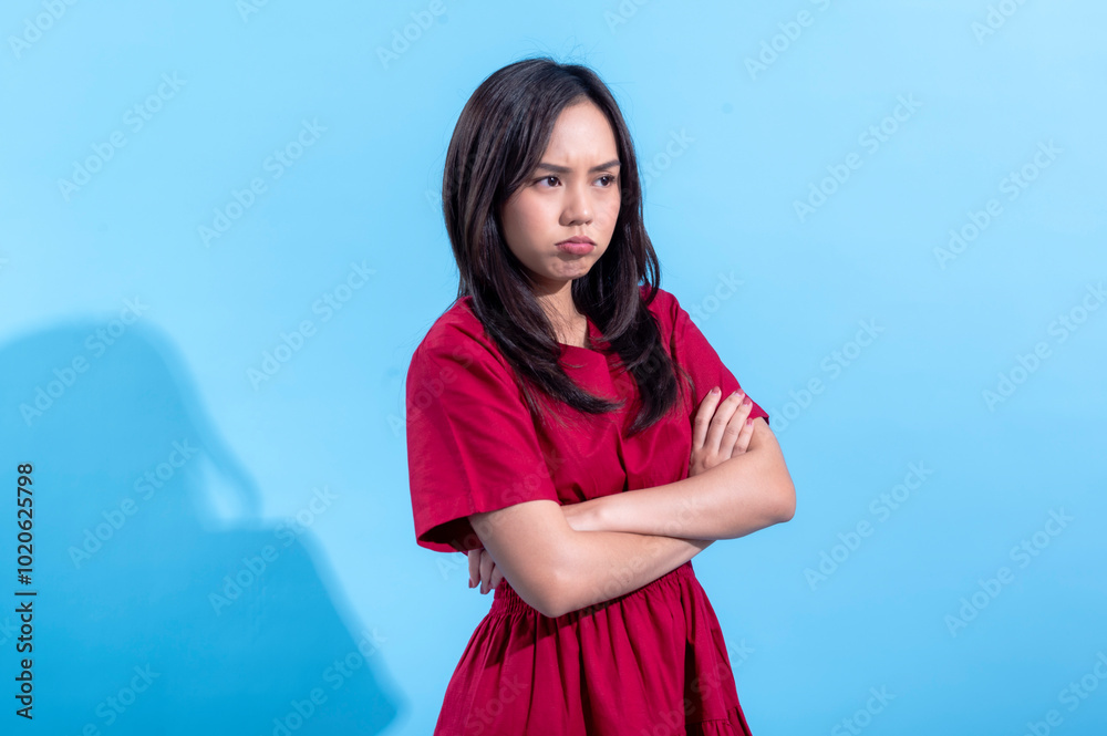 An Asian woman wearing a red dress, crossing her arms with a pouty expression, showing frustration or sulking. She stands against a light blue background, emphasizing her displeased An Asian woman wearing a red dress, crossing her arms with a pouty expression, showing frustration or sulking. She stands against a light blue background, emphasizing her displeased