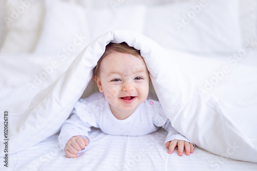 funny happy baby girl looks out from under the blanket on the bed in white clothes and smiles, a small child on a cotton bed at home woke up in the morning after sleeping