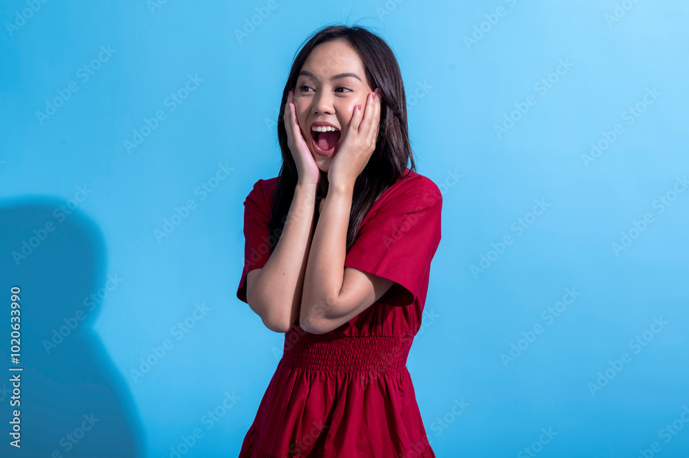 An Asian woman in a red dress looks shocked with wide eyes and both hands pressed against her cheeks. She expresses surprise and disbelief. The background is light blue An Asian woman in a red dress looks shocked with wide eyes and both hands pressed against her cheeks. She expresses surprise and disbelief. The background is light blue