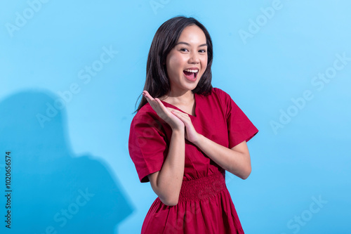 Asian woman wearing a red dress claps her hands in excitement while looking off to the side. She appears joyful and expressive against a light blue background, conveying positive energy and enthusiasm