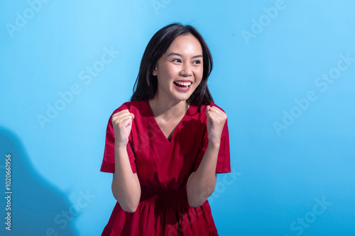 An excited Asian woman in a red dress raises both fists in the air while cheering enthusiastically. She stands against a light blue background, displaying an expression of joy and triumph photo