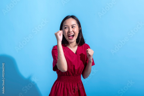 An excited Asian woman in a red dress raises both fists in the air while cheering enthusiastically. She stands against a light blue background, displaying an expression of joy and triumph photo