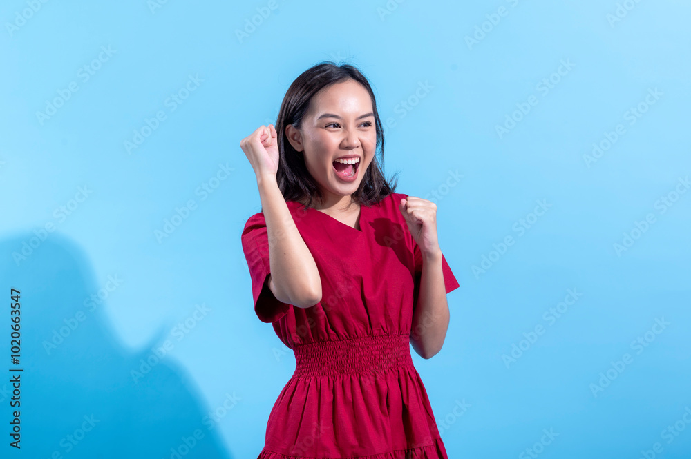 An excited Asian woman in a red dress raises both fists in the air while cheering enthusiastically. She stands against a light blue background, displaying an expression of joy and triumph An excited Asian woman in a red dress raises both fists in the air while cheering enthusiastically. She stands against a light blue background, displaying an expression of joy and triumph