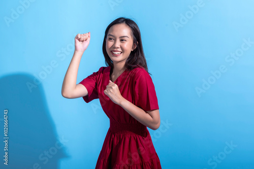 An excited Asian woman in a red dress raises both fists in the air while cheering enthusiastically. She stands against a light blue background, displaying an expression of joy and triumph photo