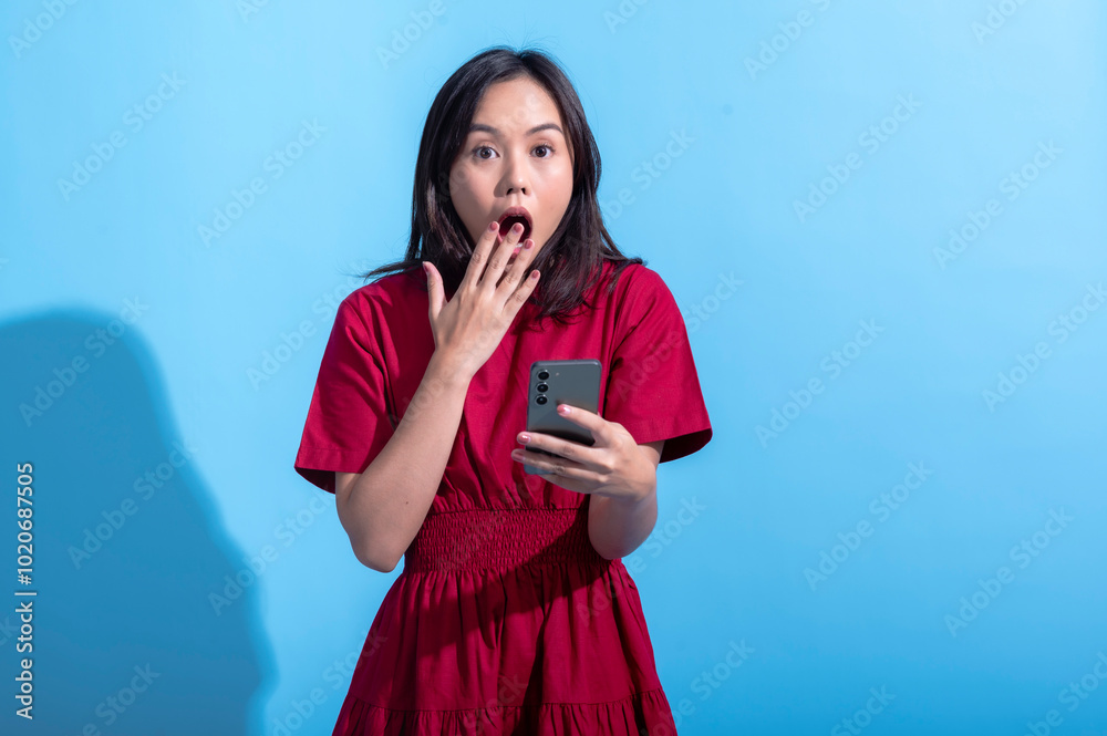 An Asian woman wearing a red dress is shocked while looking at her smartphone. Her hand covers her mouth in surprise, and she holds the phone in her other hand. The background is a light blue color An Asian woman wearing a red dress is shocked while looking at her smartphone. Her hand covers her mouth in surprise, and she holds the phone in her other hand. The background is a light blue color