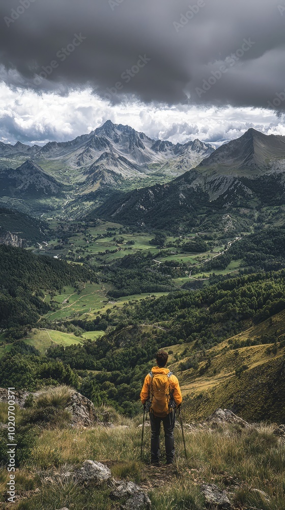 Obraz premium A hiker stands on a mountain peak, overlooking a vast valley surrounded by majestic peaks under a moody sky.