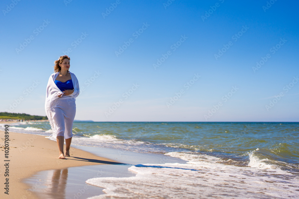 Summer vacations. Beautiful young woman in blue swimsuit covered with white scarf walking on sand by sea on beach on sunny day. Summer vacations. Beautiful young woman in blue swimsuit covered with white scarf walking on sand by sea on beach on sunny day.