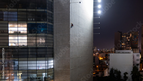 Man Hanging Up Working on the Wall of a Building Under Construction at Night with Horizontal Lights on Poles and Offices with Light