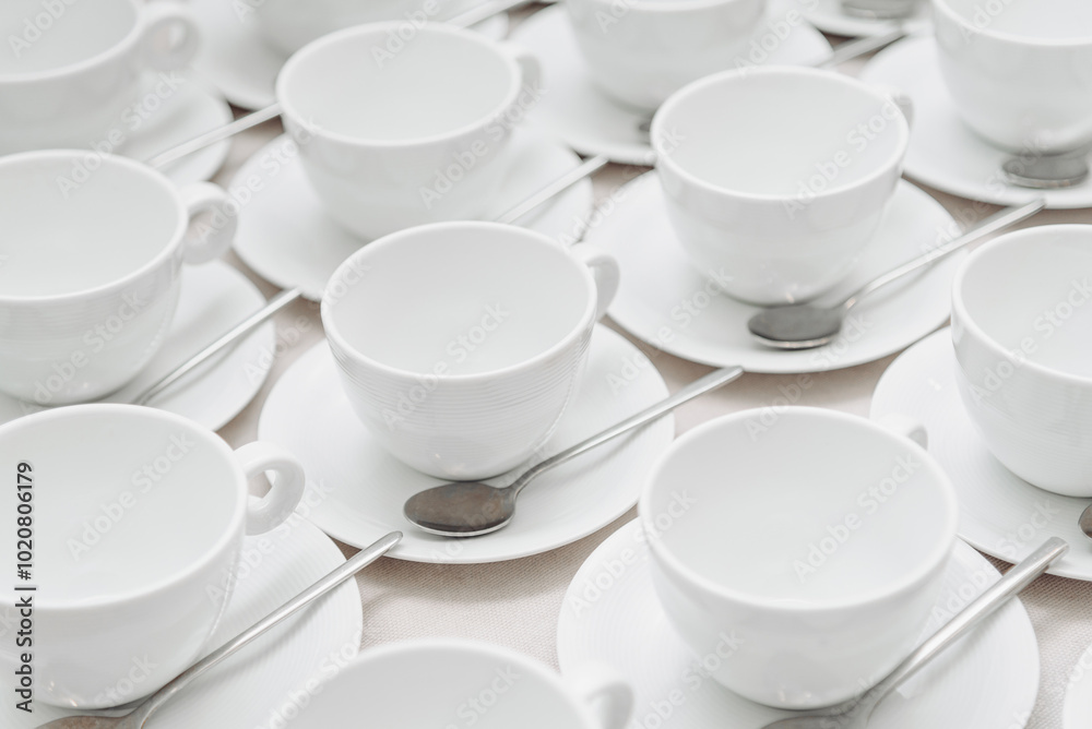 white empty porcelain cups on saucers with spoons in several rows on white tablecloth, serving tables for banquet in restaurant or cafe, pattern, background white empty porcelain cups on saucers with spoons in several rows on white tablecloth, serving tables for banquet in restaurant or cafe, pattern, background