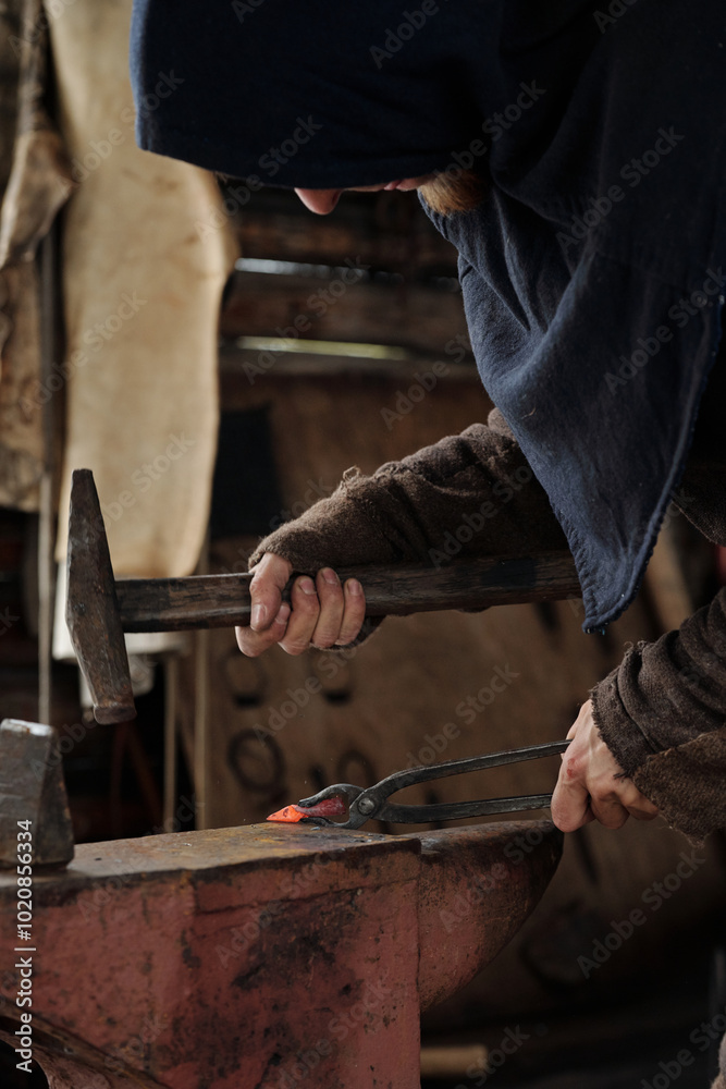 Person hammering heated metal on anvil in dimly lit workshop, wearing protective clothing, creating a traditional blacksmithing scene without modern tools Person hammering heated metal on anvil in dimly lit workshop, wearing protective clothing, creating a traditional blacksmithing scene without modern tools