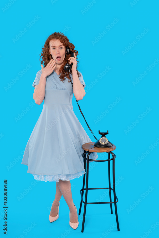A surprised woman in a blue dress talks on a vintage telephone against a bright blue background A surprised woman in a blue dress talks on a vintage telephone against a bright blue background