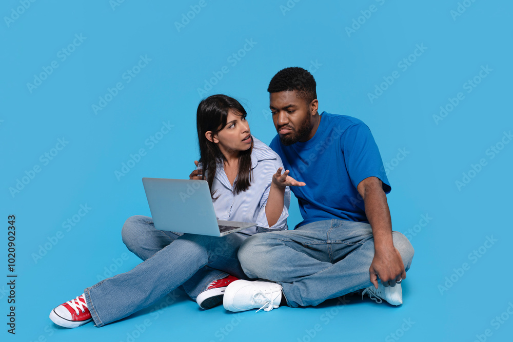 Young couple engaged in a lively discussion while using a laptop against a vibrant blue background Young couple engaged in a lively discussion while using a laptop against a vibrant blue background