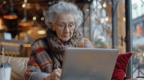 An elderly woman with a determined expression, using a laptop in a cozy cafe setting. She is wearing glasses and a warm sweater