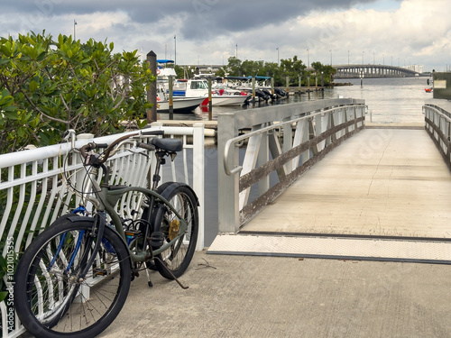 Two bicycles parked together by entrance ramp at a riverside marina in Punta Gorda, Florida, with a view of the northbound US 41 traffic bridge across Peace River, on a cloudy day in southwest Florida