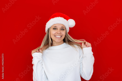 A happy young charming smiling blonde woman in a Santa hat and a white sweatshirt with blank copy space holding her strands of hair on a red color background. Christmas and New Year concept. Mockup