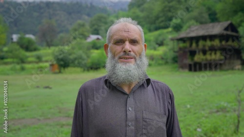 A close up shot of a happy Indian Kashmiri bearded old or aged Muslim man or male in ethnic attire standing outdoors on a green meadow with scenic old village houses facing the camera and smiling