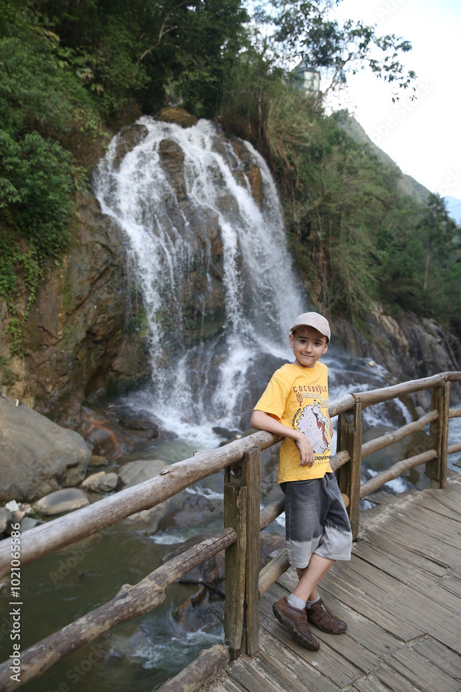 Obraz premium Tourist child, cute kid, fashion boy in Cat Cat village. Landmark of Sa Pa city, northern Vietnam. Vietnamese nature, beautiful waterfall, scenery, landscape. Wooden bridge. Tourism, travel in Sa Pa