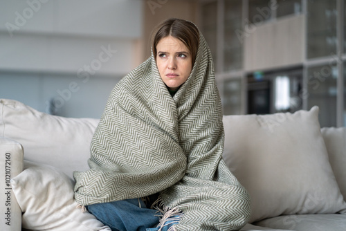 Offended lonely woman sits motionless on sofa, wrapped in blanket, overwhelmed by depression and sadness. Female with emotional exhaustion, autumn blues, mental health problem, insomnia and illness