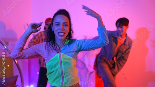 Attractive happy dancer looking at camera while smart woman moving at rhyme with neon light. Professional hispanic performer break dancing while wearing colorful cloth with diverse friend. Regalement.