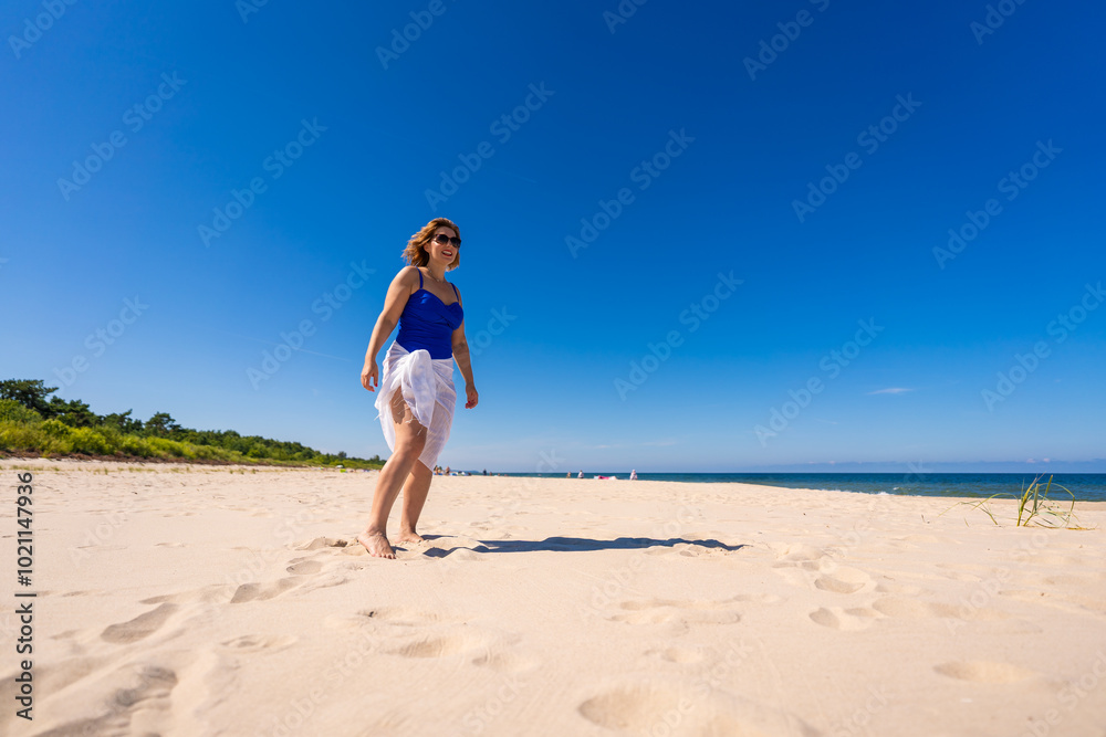 Summer vacation. Beautiful young woman in one-piece blue swimsuit and white scarf and sunglasses walking on sandy beach on beautiful sunny day. Sunbathing. Summer vacation. Beautiful young woman in one-piece blue swimsuit and white scarf and sunglasses walking on sandy beach on beautiful sunny day. Sunbathing.
