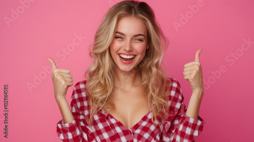 A cheerful young woman with blonde hair gives a thumbs up gesture while smiling broadly against a pink background. photo