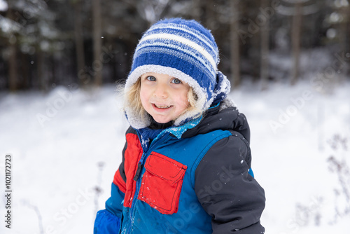 Happy young boy is standing in a snowy forest wearing a warm winter coat and hat. Selective focus