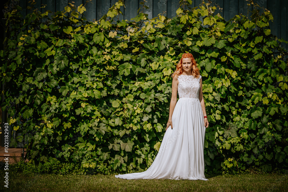Bride in a white lace dress stands against a lush green leafy background outdoors, with sunlight highlighting her red hair.. Bride in a white lace dress stands against a lush green leafy background outdoors, with sunlight highlighting her red hair..
