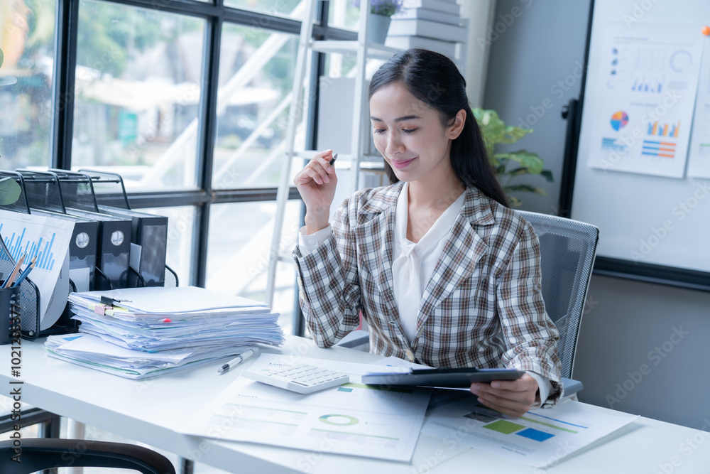 Accountant is reviewing financial statements and calculating taxes while working in her office. She is surrounded by paperwork and using a calculator