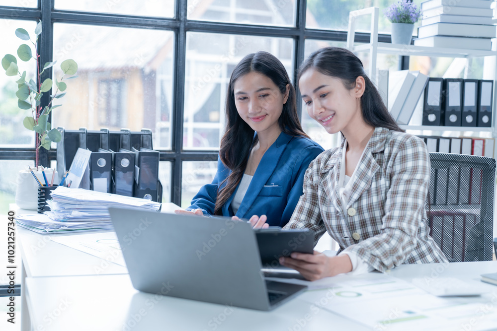 Two young businesswomen collaborate in a modern office, looking at a laptop with charts and documents. Natural light illuminates them as they smile confidently Two young businesswomen collaborate in a modern office, looking at a laptop with charts and documents. Natural light illuminates them as they smile confidently