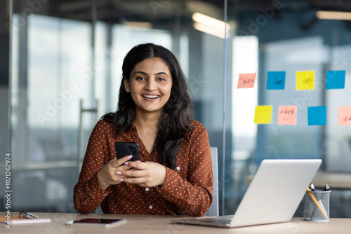 Smiling young professional woman holding smartphone, seated at office desk with laptop, notes, and sticky notes in background, conveying happiness and productivity. Setting suggests modern workspace.