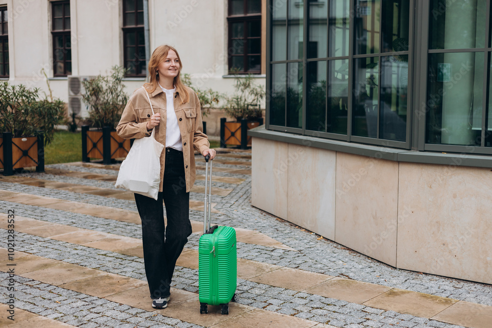 Cheerful young woman in casual outfit walking on the city street with green luggage. 30s Woman dragging suitcase on urban background. Travel Fashion. Full body