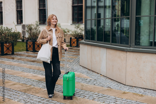 Obraz na płótnie Cheerful young woman in casual outfit walking on the city street with green luggage. 30s Woman dragging suitcase on urban background. Travel Fashion. Full body w salonie