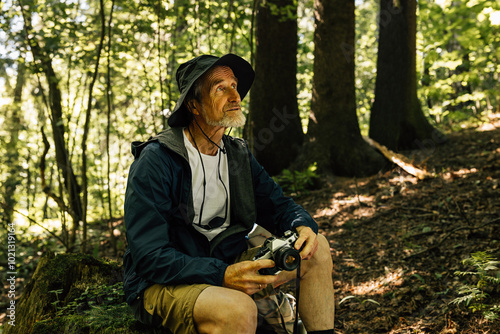 Senior male sitting in the forest and holding a film camera. Mature male in a hat and hiking clothes taking a break during a forest walk.