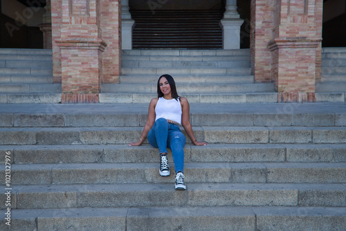 Moroccan woman, young, beautiful, brunette, with white top and jeans, relaxed and calm looking at the camera, sitting on some steps. Concept of ethnicity, diversity, femininity, beauty.