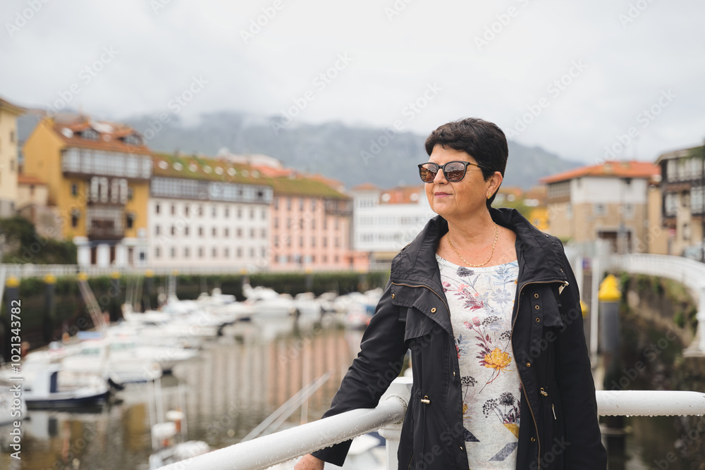 Fototapeta premium Senior woman wearing sunglasses is standing on a bridge and enjoying the view of llanes harbor in asturias, spain