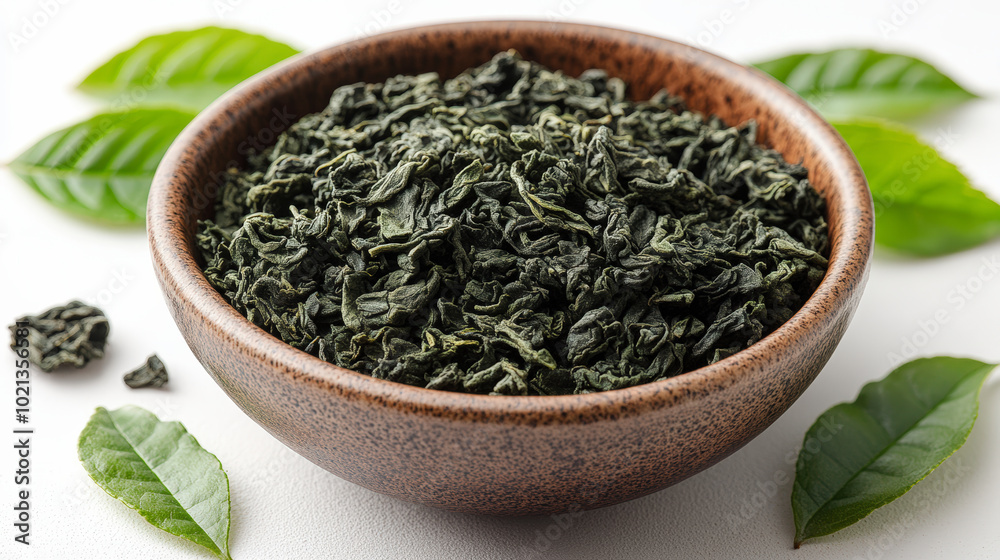 A rustic bowl filled with dried tea leaves is surrounded by fresh green leaves on a white surface, creating a refreshing natural scene. A rustic bowl filled with dried tea leaves is surrounded by fresh green leaves on a white surface, creating a refreshing natural scene.