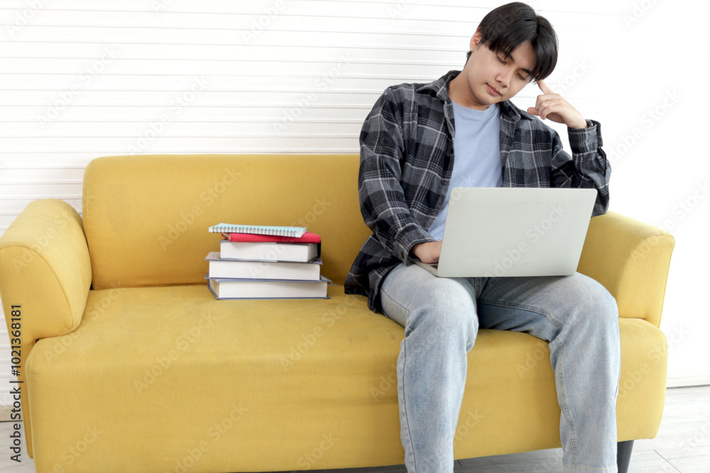 Stress Asian high school boy look on laptop computer while sits on sofa with pile of books. University male student study online. College pupil learn tutor video to prepare exam. Senior education.