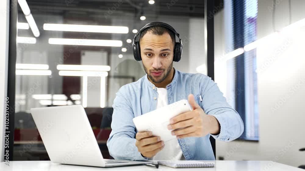 Young adult african american businessman in wireless headphones plays computer game on digital tablet while sitting at desk at workplace in business office. Excited male engrossed in interesting game Young adult african american businessman in wireless headphones plays computer game on digital tablet while sitting at desk at workplace in business office. Excited male engrossed in interesting game