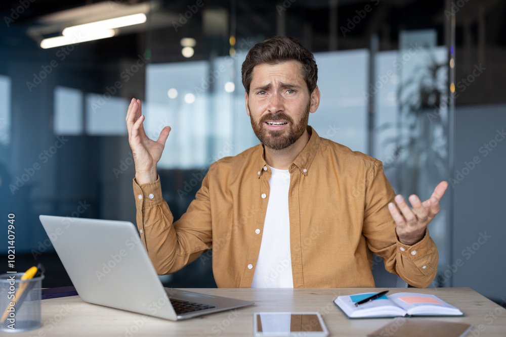 Frustrated businessman gestures at desk with laptop, notebook, phone, suggesting tech issue or confusion in office. Uncertainty and stress in workplace captured in moment. Frustrated businessman gestures at desk with laptop, notebook, phone, suggesting tech issue or confusion in office. Uncertainty and stress in workplace captured in moment.