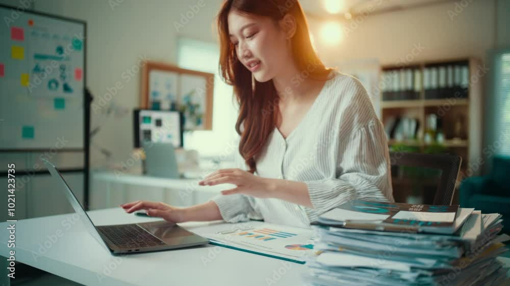 Asian accountant woman is working hard at her desk in the office. She is surrounded by paperwork and is typing on her laptop Asian accountant woman is working hard at her desk in the office. She is surrounded by paperwork and is typing on her laptop
