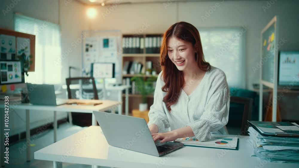 Young businesswoman is analyzing charts and working on a laptop at her office desk. She is surrounded by paperwork and office supplies Young businesswoman is analyzing charts and working on a laptop at her office desk. She is surrounded by paperwork and office supplies