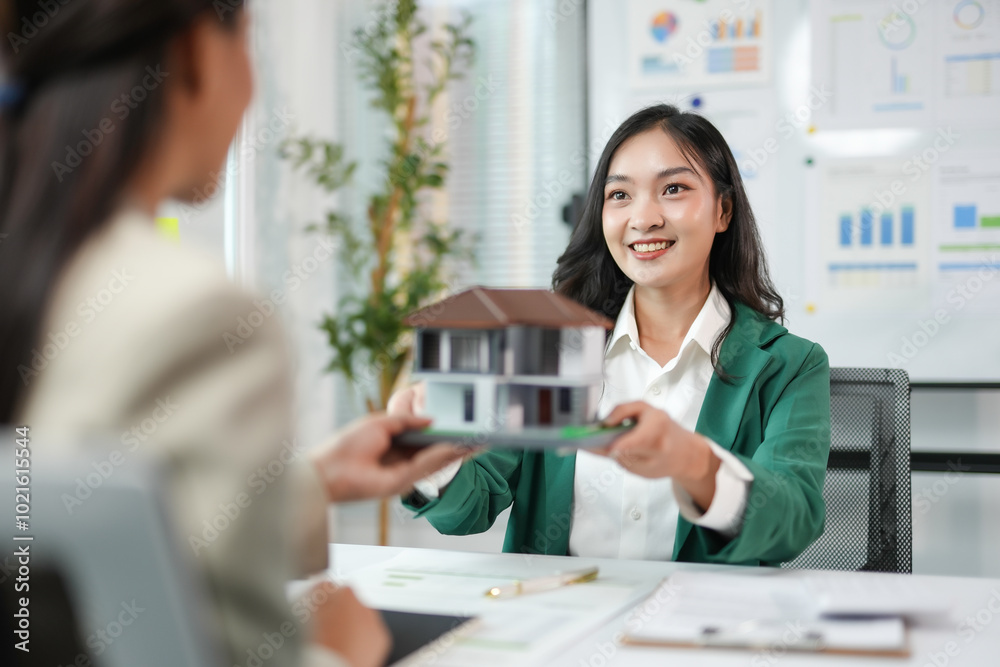 Smiling real estate agent holding a model house, while giving it to a client during a meeting Smiling real estate agent holding a model house, while giving it to a client during a meeting